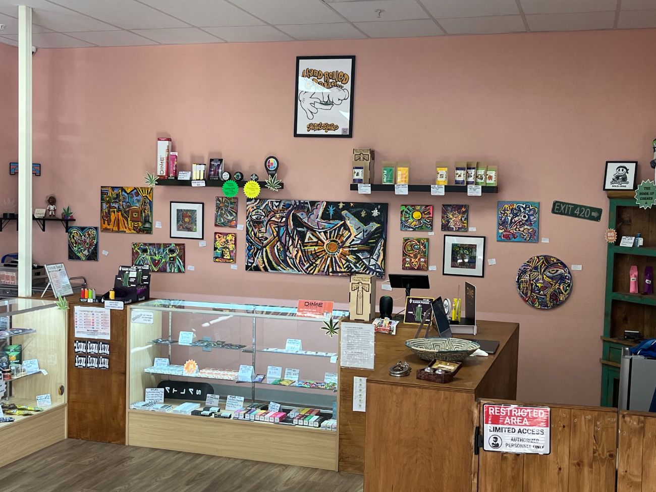 The main counter at TOKE It Up: glass display cases, a wooden register counter, and a rose-pink wall hung with paintings and shelves.