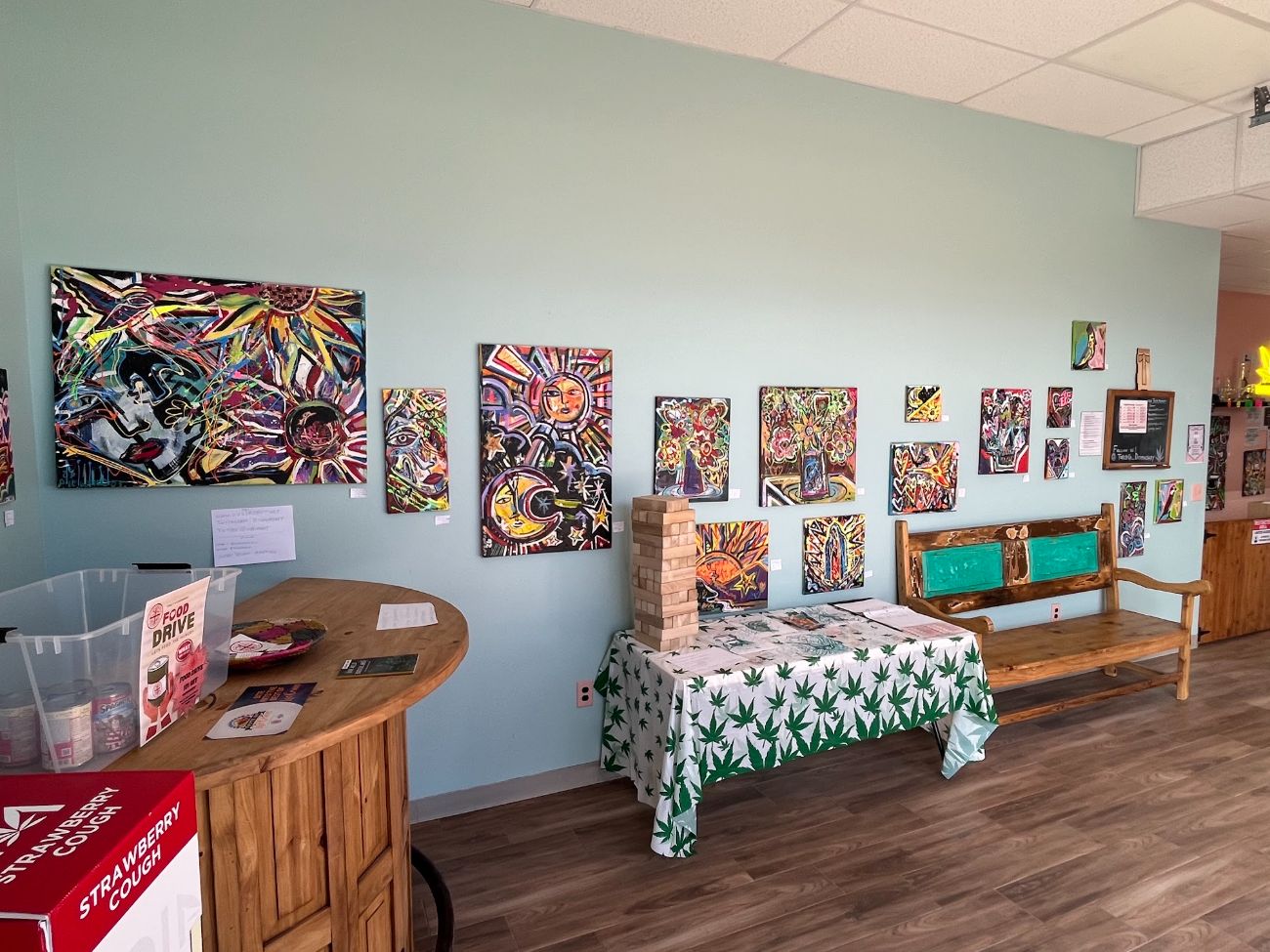 The mint-green side of the shop: a wooden bench, a table with a cannabis-leaf cloth and a Jenga tower, and a wall of paintings.