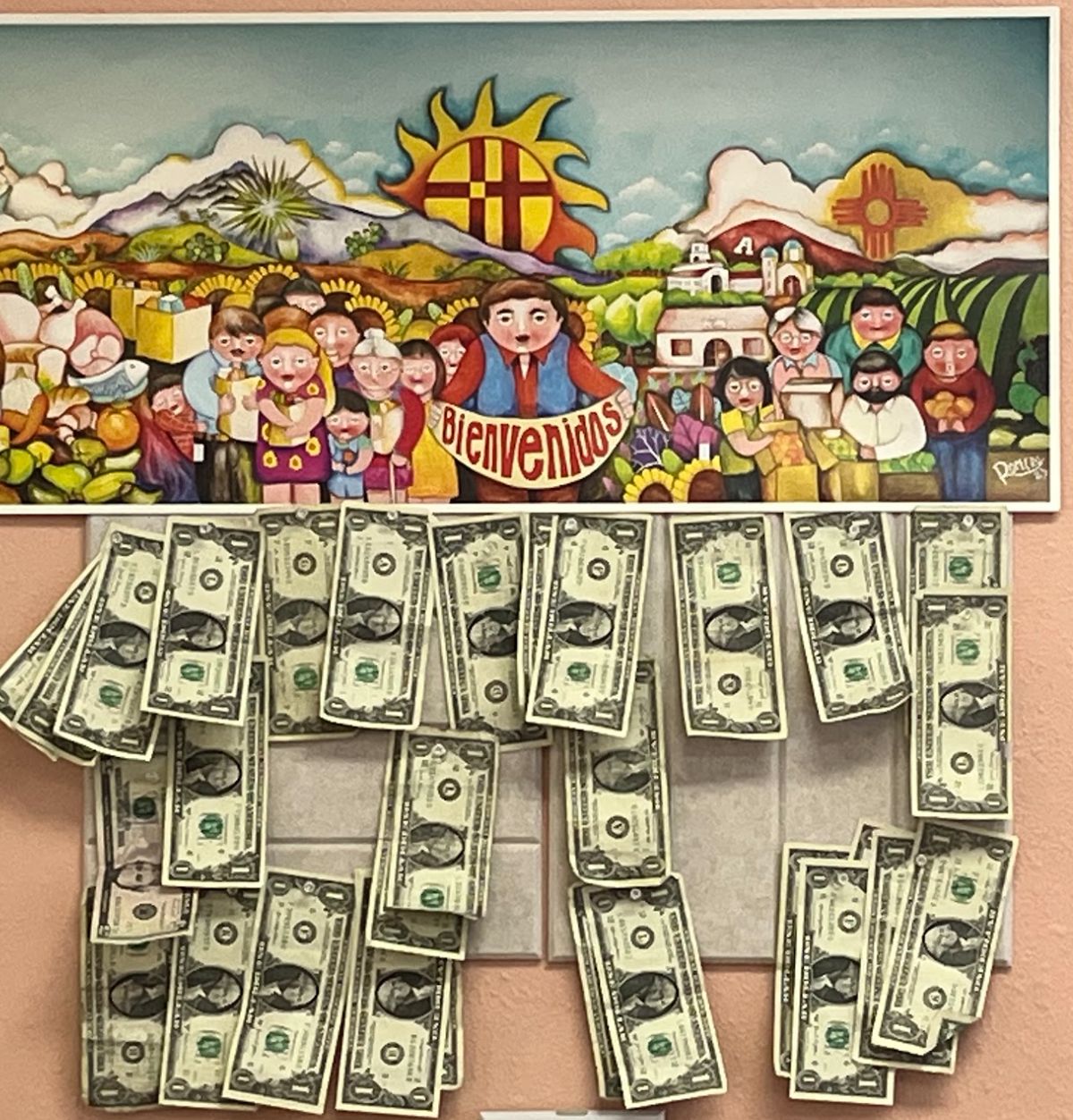 Inside TOKE It Up: a colourful Casa De Peregrinos Food Program poster reading Bienvenidos, with dozens of dollar bills pinned to the wall beneath it.